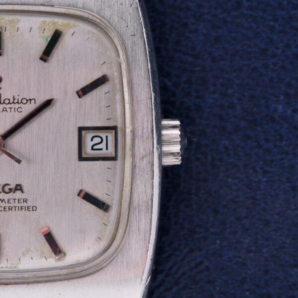 Close-up of an Omega Constellation TV Case Certified Chronometer watch face with a square design, time at 10:15, and date on 21. Hour markers pop against a textured background on a dark surface.