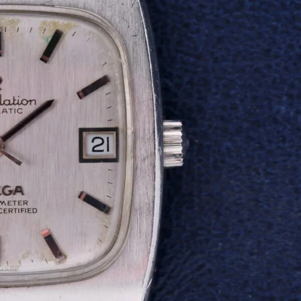 Close-up of an Omega Constellation TV Case Certified Chronometer watch face with a square design, time at 10:15, and date on 21. Hour markers pop against a textured background on a dark surface.