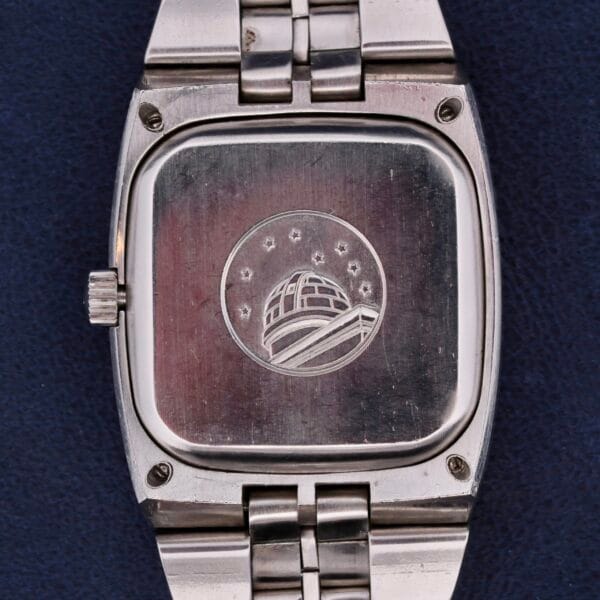 The image displays an Omega Constellation TV Case Certified Chronometer, featuring its silver back engraved with a dome and stars logo. It has a sleek metal link strap and is elegantly placed on a dark blue surface.