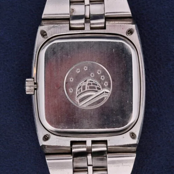 The image displays an Omega Constellation TV Case Certified Chronometer, featuring its silver back engraved with a dome and stars logo. It has a sleek metal link strap and is elegantly placed on a dark blue surface.
