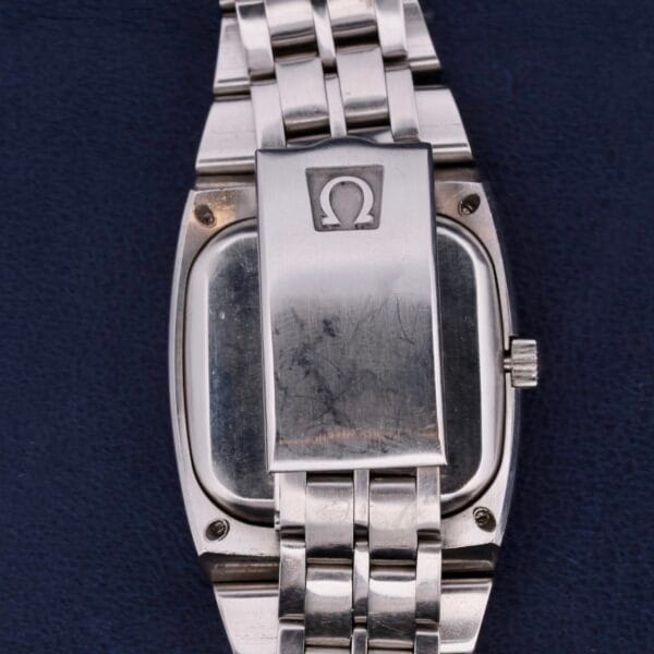 A silver wristwatch with a metal link band lies on a dark blue surface. Its clasp features the Omega logo, an uppercase Greek letter omega (Ω), identifying it as an elegant Omega Constellation TV Case Certified Chronometer.