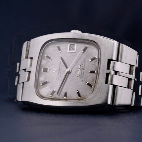 The Omega Constellation TV Case Certified Chronometer features a rectangular silver face with a textured dial, contrasting hour markers, hands, and a date display at the top right, set on a metal link band.