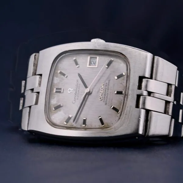 The Omega Constellation TV Case Certified Chronometer features a rectangular silver face with a textured dial, contrasting hour markers, hands, and a date display at the top right, set on a metal link band.
