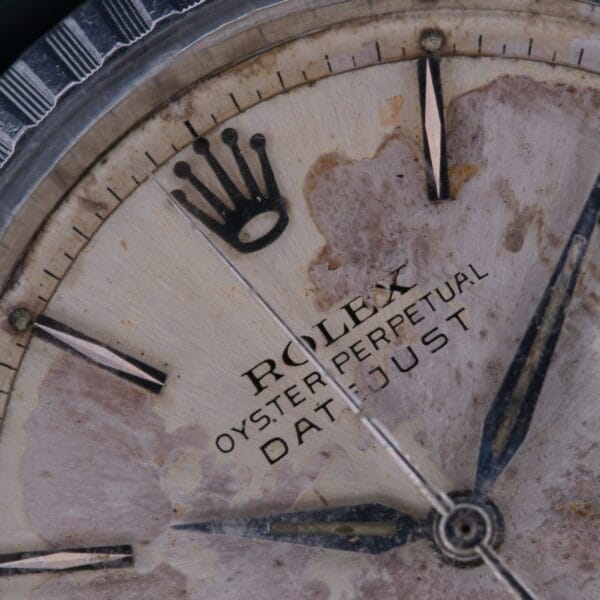 A close-up of the aged Rolex Datejust 1603 Pie-Pan Dial reveals its storied past, with noticeable wear and discoloration. The black crown logo and dark hour markers stand boldly against the beige, spotted background, showcasing the iconic Oyster Perpetual design.