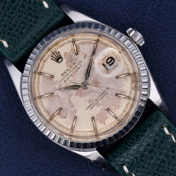 The Rolex Datejust 1603 Pie-Pan Dial features a silver bezel, green leather strap, weathered dial, and date display at 3 oclock, with black hour markers and hands on a blue textured background.