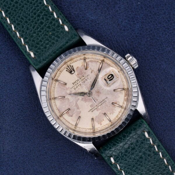 A vintage Rolex Datejust 1603 Pie-Pan Dial wristwatch, with a worn off-white face featuring a date window and silver hour markers in a metal casing. It has a textured green leather strap and sits elegantly on a dark blue surface.