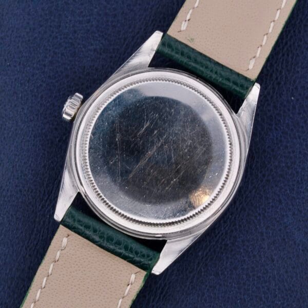 A scratched Rolex Datejust 1603 Pie-Pan Dial case, lacking a face, is attached to a two-toned leather strap in cream and dark green segments on a textured dark blue surface.