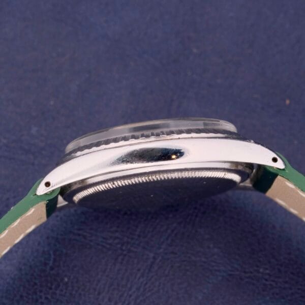 A close-up side view of the Rolex Datejust 1603 Pie-Pan Dial watch with a silver case and green leather strap against a dark blue textured background, partially showcasing the bezel and edge details.