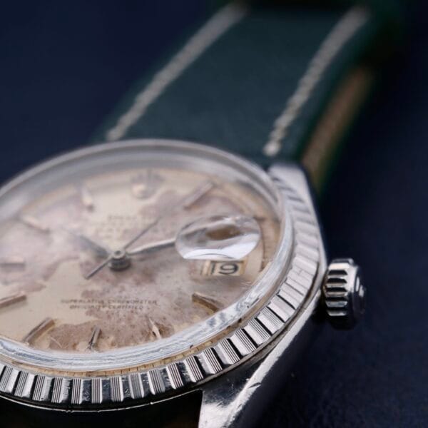 Close-up of a vintage Rolex Datejust 1603 Pie-Pan Dial wristwatch with a green leather strap and an aged, off-white dial, featuring a magnified date window and fluted bezel against a dark blue background.