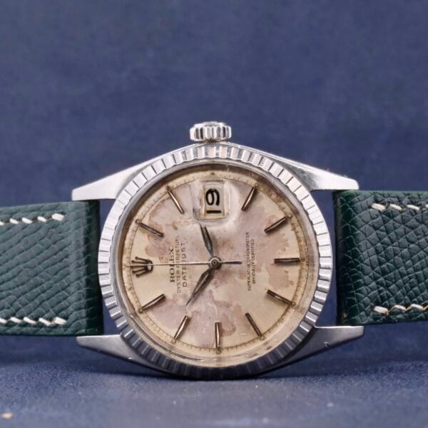 The Rolex Datejust 1603 Pie-Pan Dial is a vintage wristwatch with a worn beige dial, baton hour markers, and day-date function. It features a silver bezel and a textured green leather strap against a plain blue background.