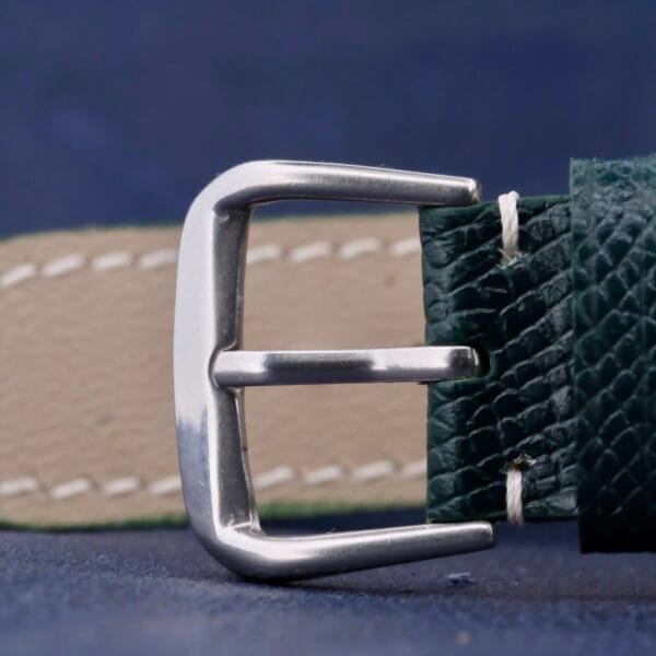 Close-up of a Rolex Datejust 1603 Pie-Pan Dial-inspired silver metallic belt buckle on a dark green textured leather strap with beige stitching, elegantly arranged on a dark blue surface.