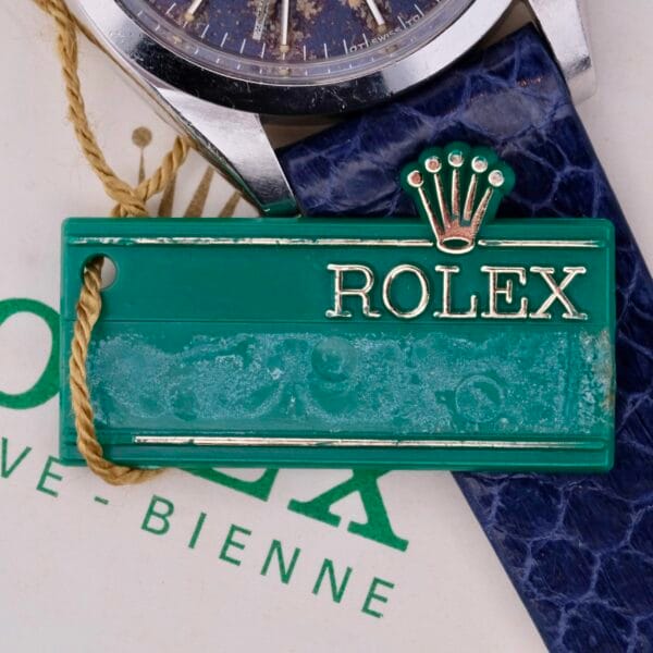 Close-up of the iconic Rolex green tag attached to a blue strap with a string, featuring the logo and name. Above the tag, part of the elegant Rolex Oyster Perpetual Date 1500 Blue Dial watch face is visible, hinting at its timeless allure.