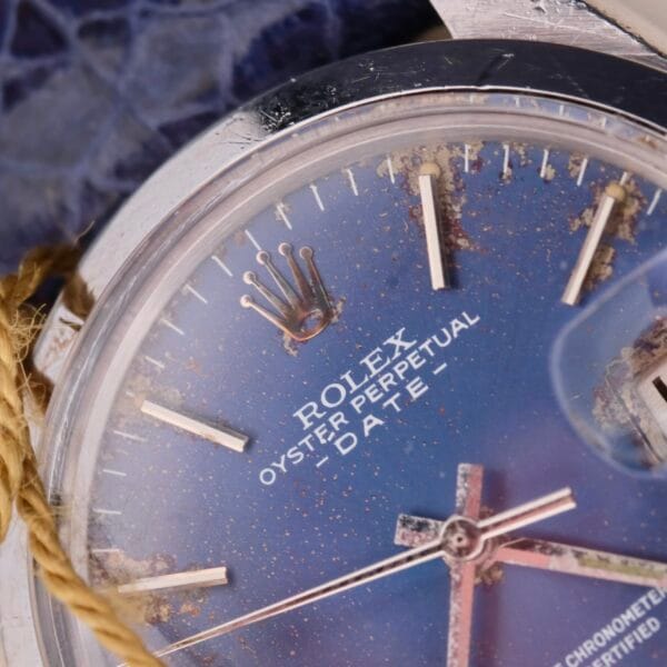 The Rolex Oyster Perpetual Date 1500 Blue Dial watch showcases a close-up of its blue face. The fading and speckling on the dial are signs of wear over time, while the iconic Rolex logo and text remain prominently beneath the glass.