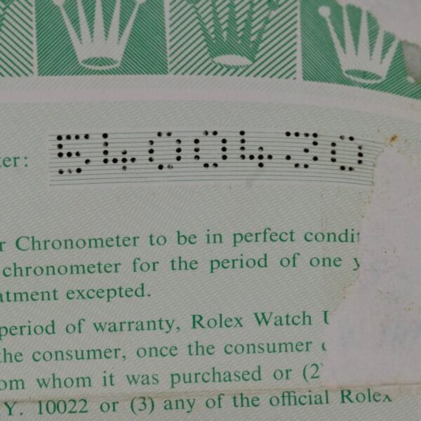 A close-up shows a document with perforated serial number 5600430, set against a luxury-inspired green pattern with partial crown logos reminiscent of the Rolex Oyster Perpetual Date 1500 Blue Dial. Text is partially visible below the serial number.