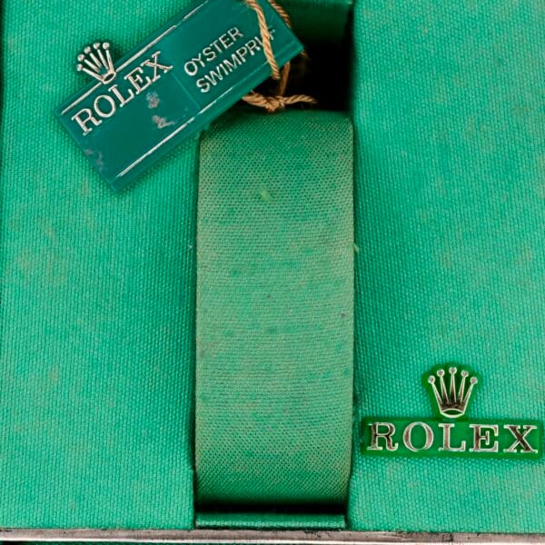 The green watch box features a central green cushion, a Rolex Oyster Swimpruf tag, and Rolex with the crown logo on the side. Elegantly crafted to hold your Rolex Oyster Perpetual Date 1500 Blue Dial securely.
