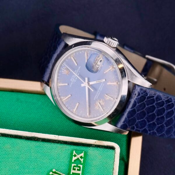 The Rolex Oyster Perpetual Date 1500 Blue Dial, with a blue dial and leather strap, a silver casing, and a date display under a magnifier, is positioned next to a green textured box partially revealing gold text.