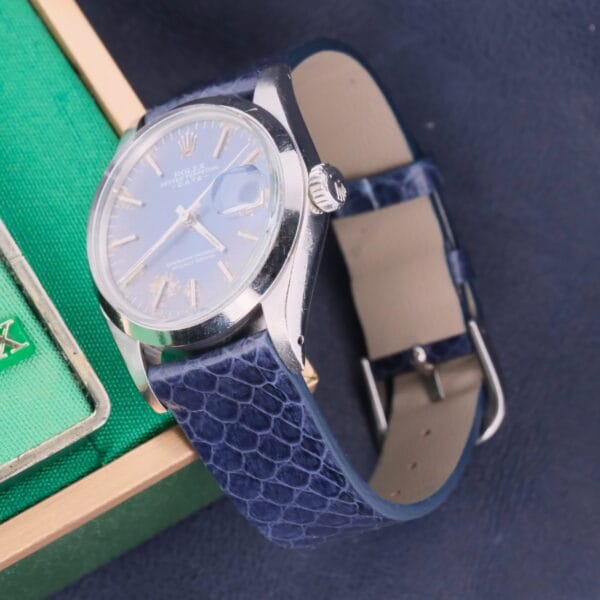A Rolex Oyster Perpetual Date 1500 with a blue leather strap and white dial featuring a date display sits on a dark surface, accompanied by a partially visible green box.