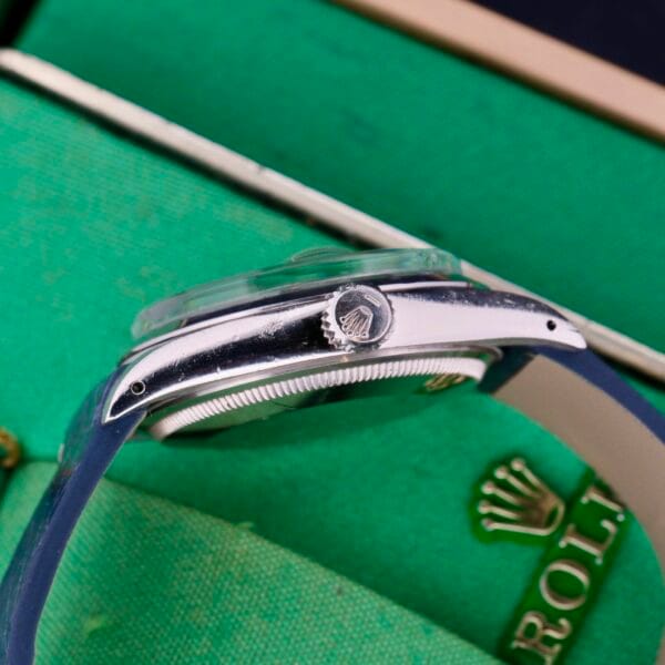 A close-up side view of a luxury Rolex Oyster Perpetual Date 1500 Blue Dial wristwatch with a silver case and blue strap. The watch crown displays an embossed logo, while a green box in the background partially shows the iconic gold ROLEX logo.