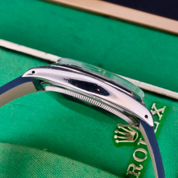 Side view of the Rolex Oyster Perpetual Date 1500 Blue Dial, showcasing a stainless steel case and blue strap. It rests elegantly on a green surface with part of a logo in the background.