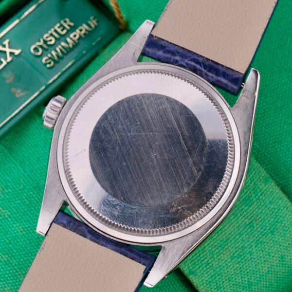 The Rolex Oyster Perpetual Date 1500 Blue Dial silver watch case, without its face, sits on a green table. It has a blue and beige strap. A green and white label is partially visible in the background.