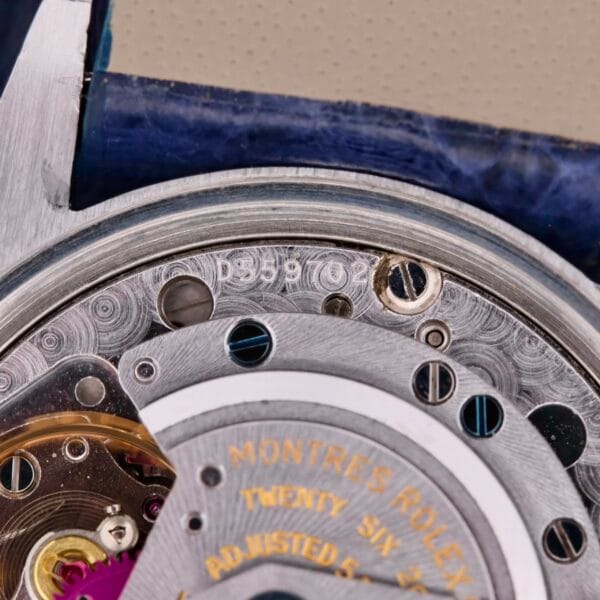 Close-up of a Rolex Oyster Perpetual Date 1500 Blue Dial showing intricate metallic gears and components, with engraved numbers and Rolex branding. The watch casing edge is styled in blue.