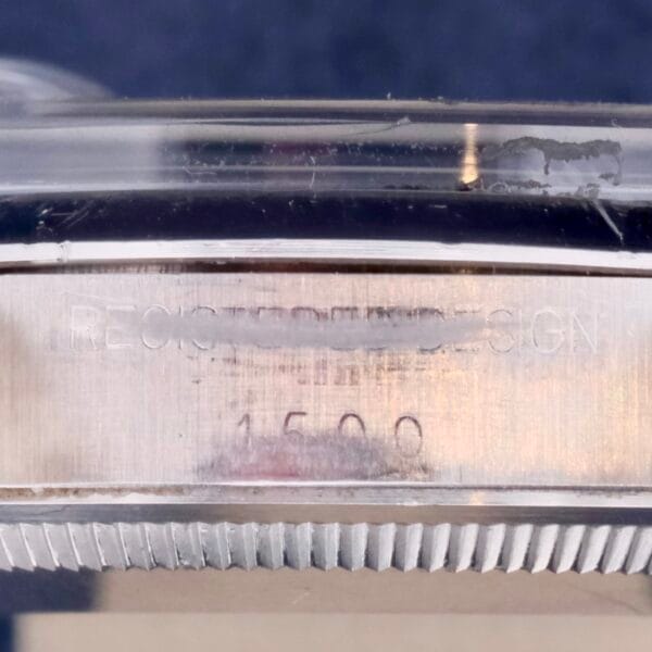 A close-up of a metallic surface shows engraved text. The blurred upper text resembles PRECISION DESIGNS, akin to the Rolex Oyster Perpetual Date 1500 Blue Dial, with 1500 clearly visible below. The reflective finish and ridged edge highlight its elegant design.