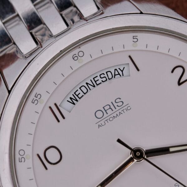 Close-up of the Oris XXL Full Day Automatic silver watch displaying Wednesday, with black hour markers at 10, 11, and 12, sleek metallic hands, and a metal link strap.