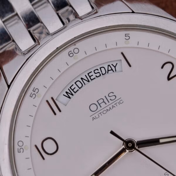 Close-up of the Oris XXL Full Day Automatic silver watch displaying Wednesday, with black hour markers at 10, 11, and 12, sleek metallic hands, and a metal link strap.
