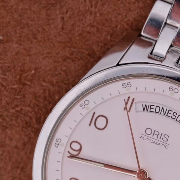 A close-up of the silver Oris XXL Full Day Automatic showcases its white dial, displaying Wednesday and numbers 10, 11, and 12 in a minimalist font. The background is a textured brown surface.