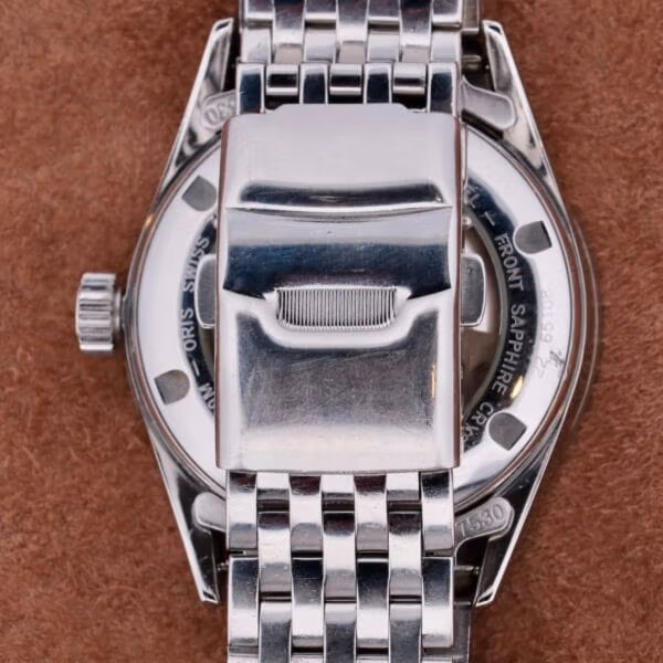 The rear of the Oris XXL Full Day Automatic in stainless steel, with its intricate metal band, highlights an elegant clasp and detailed back against a textured brown surface.
