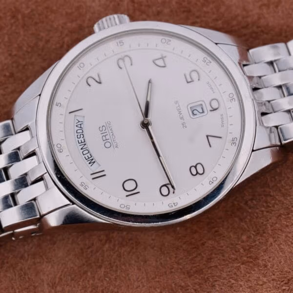 The Oris XXL Full Day Automatic wristwatch features a white dial with Arabic numerals, displaying Wednesday the 10th, and metallic hour and minute hands. It includes a linked metal bracelet on a brown textured surface.