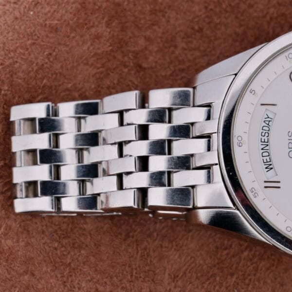 Close-up of a sleek Oris XXL Full Day Automatic wristwatch with a metal link band on a weathered brown background. The watch face is partially visible, displaying Wednesday and part of the logo.