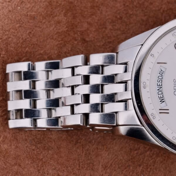 Close-up of a sleek Oris XXL Full Day Automatic wristwatch with a metal link band on a weathered brown background. The watch face is partially visible, displaying Wednesday and part of the logo.