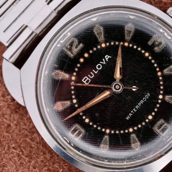 Close-up of a vintage Bulova Seabee Charcoal Waffle Dial wristwatch featuring a charcoal waffle face, silver numbers, gold hands, and a silver metal bezel and band. The dial reads WATERPROOF. Its resting on a brown textured surface.