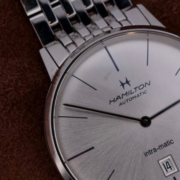 Close-up of a Hamilton Intramatic Date Stainless Steel automatic wristwatch, featuring a silver face, black hour markers, and a date window at 6 oclock. The watch has a stainless steel bracelet on a textured brown background.