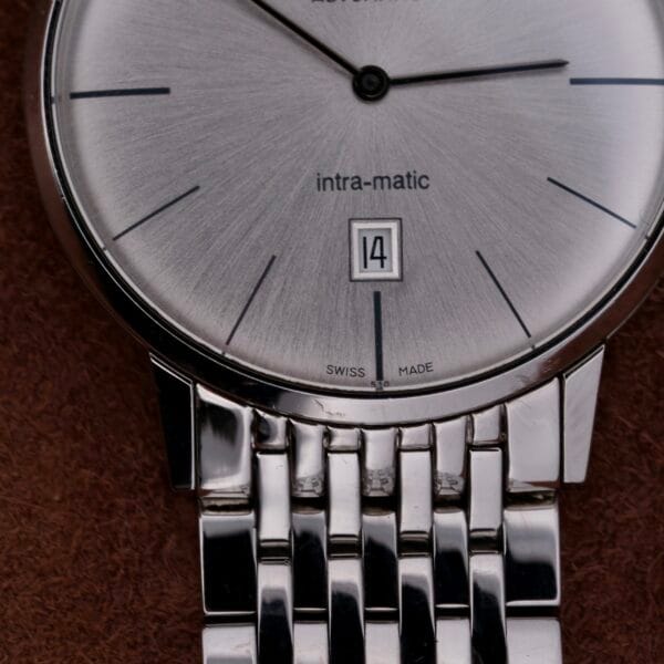A close-up of the Hamilton Intramatic Date Stainless Steel wristwatch reveals its minimalist design, with slender black hour markers, intra-matic text, and a date window displaying 14. The polished metal band with a linked look is elegantly placed on a brown surface.