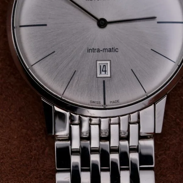 A close-up of the Hamilton Intramatic Date Stainless Steel wristwatch reveals its minimalist design, with slender black hour markers, intra-matic text, and a date window displaying 14. The polished metal band with a linked look is elegantly placed on a brown surface.