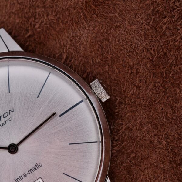 Close-up of a Hamilton Intramatic Date Stainless Steel wristwatch on a brown textured surface, highlighting its sleek circular face with minimalist hour markers, date window, and visible crown for an elegant design.