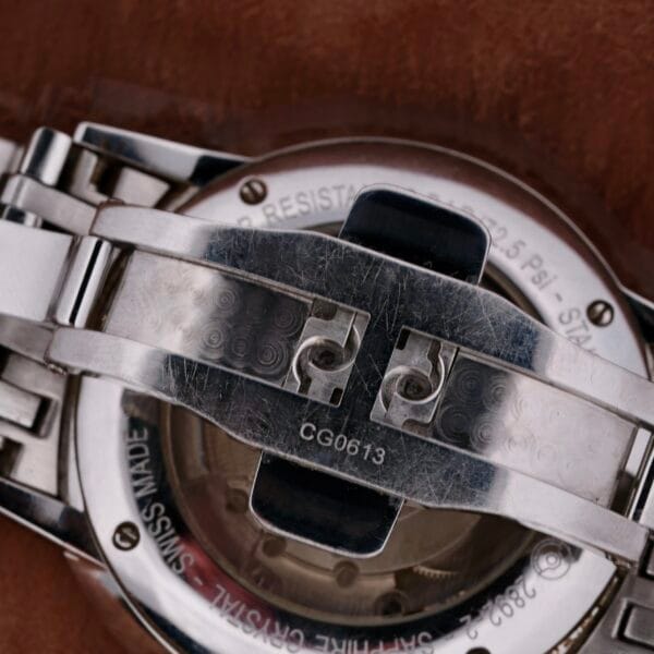 Close-up of the Hamilton Intramatic Date Stainless Steel watchs back reveals a visible folding clasp and minor scratches. Engraved with SWISS and serial number CG0613, this elegant piece rests on a rich brown leather surface, exuding timeless sophistication.