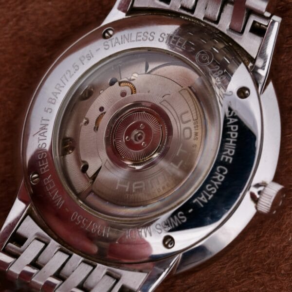 Close-up of the Hamilton Intramatic Date Stainless Steel watch back, displaying its automatic movement. The transparent case back reveals intricate gears with water resistant 5 bar, stainless steel, and sapphire crystal markings. The strap is made from sleek metal.