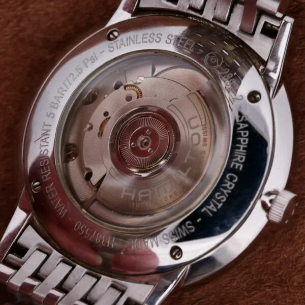Close-up of the Hamilton Intramatic Date Stainless Steel watch back, displaying its automatic movement. The transparent case back reveals intricate gears with water resistant 5 bar, stainless steel, and sapphire crystal markings. The strap is made from sleek metal.