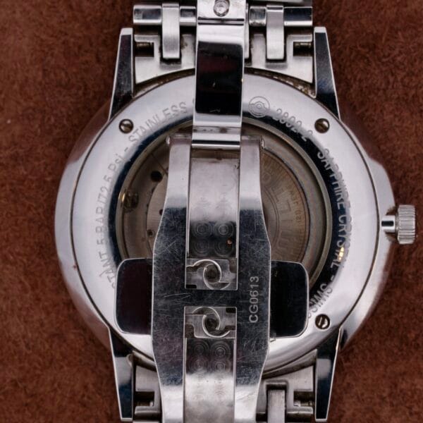 Close-up of the back of a Hamilton Intramatic Date Stainless Steel wristwatch on a brown surface, highlighting the engraved metal clasp and visible inscriptions with material and model details.