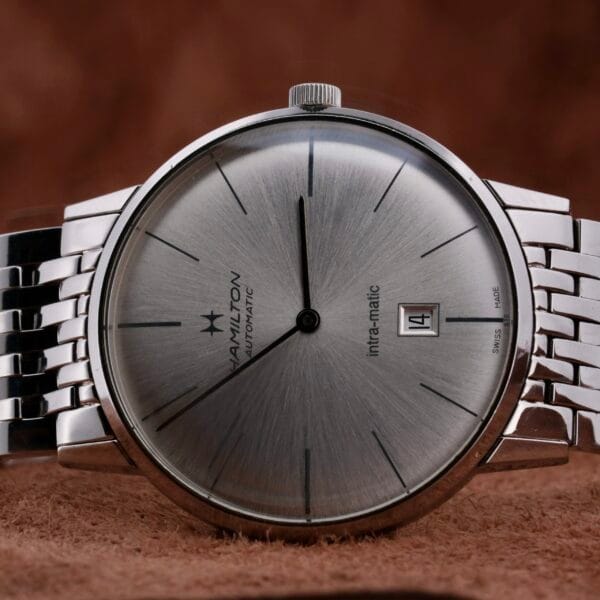 A Hamilton Intramatic Date Stainless Steel wristwatch is placed on a brown textured surface, showcasing its date set to the 14th and simple hour markers on the dial.