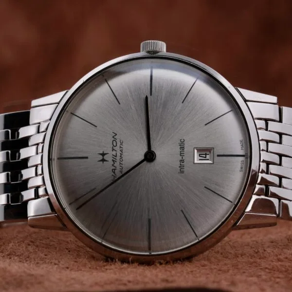 A Hamilton Intramatic Date Stainless Steel wristwatch is placed on a brown textured surface, showcasing its date set to the 14th and simple hour markers on the dial.