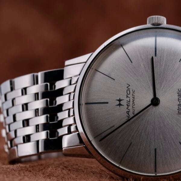 Close-up of a Hamilton Intramatic Date Stainless Steel wristwatch with a metallic bracelet, showcasing slim black hands and hour markers. The background is a soft, brown textured surface.