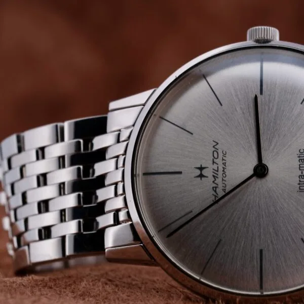 Close-up of a Hamilton Intramatic Date Stainless Steel wristwatch with a metallic bracelet, showcasing slim black hands and hour markers. The background is a soft, brown textured surface.