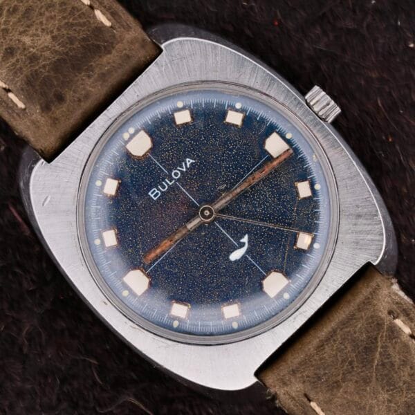 The Bulova Sea King Blue Cross Hair Dial is a vintage wristwatch with a rectangular silver case, a starry blue speckled face, square hour markers, and a small second hand subdial. Its worn brown leather strap enhances its timeless elegance and charm.