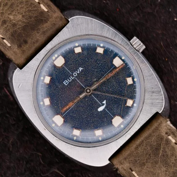 The Bulova Sea King Blue Cross Hair Dial is a vintage wristwatch with a rectangular silver case, a starry blue speckled face, square hour markers, and a small second hand subdial. Its worn brown leather strap enhances its timeless elegance and charm.