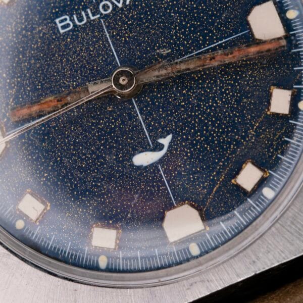 Close-up of a Bulova Sea King Blue Cross Hair Dial watch, featuring a starry night sky design on the dial with a small white whale figure. The square hour markers and hands are elegantly visible, enclosed in a sleek, round metal case.