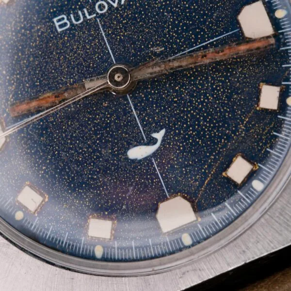 Close-up of a Bulova Sea King Blue Cross Hair Dial watch, featuring a starry night sky design on the dial with a small white whale figure. The square hour markers and hands are elegantly visible, enclosed in a sleek, round metal case.